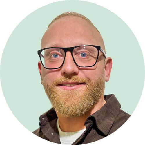 a smiling, bald man with a full blond beard and glasses, wearing a dark brown collared shirt, is pictured from the chest up against a light green background inside a white circle.
