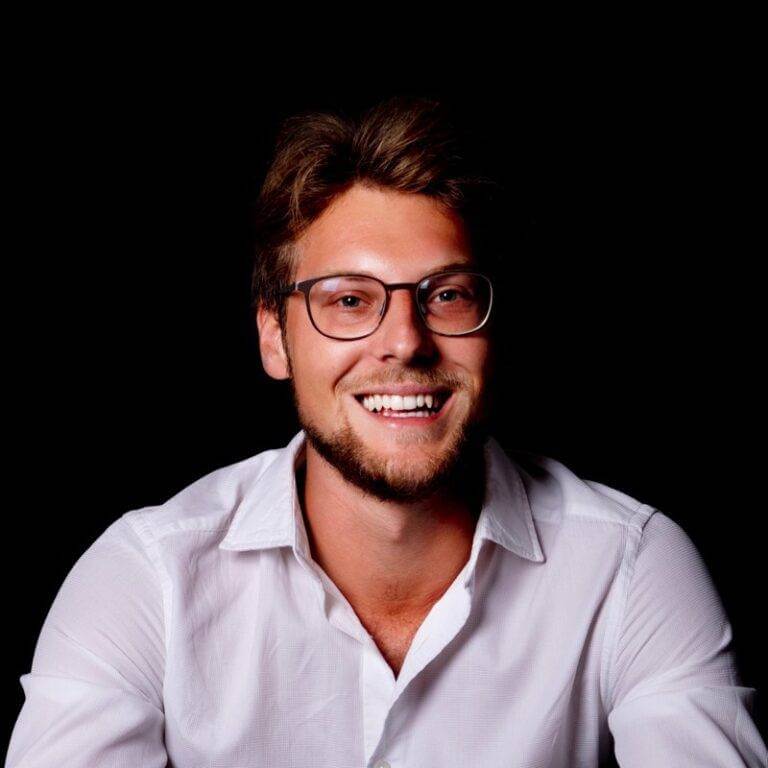 a smiling man with a beard and glasses is pictured from the chest up against a black background. he is wearing a white dress shirt.